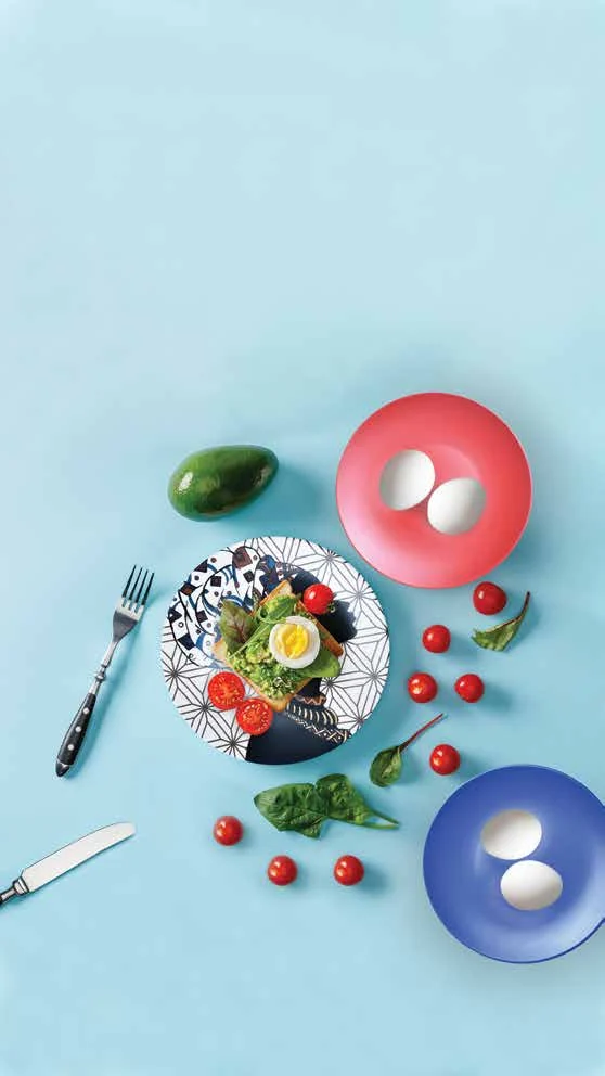 Styled breakfast scene on a pale blue background with patterned plates, eggs and cutlery.